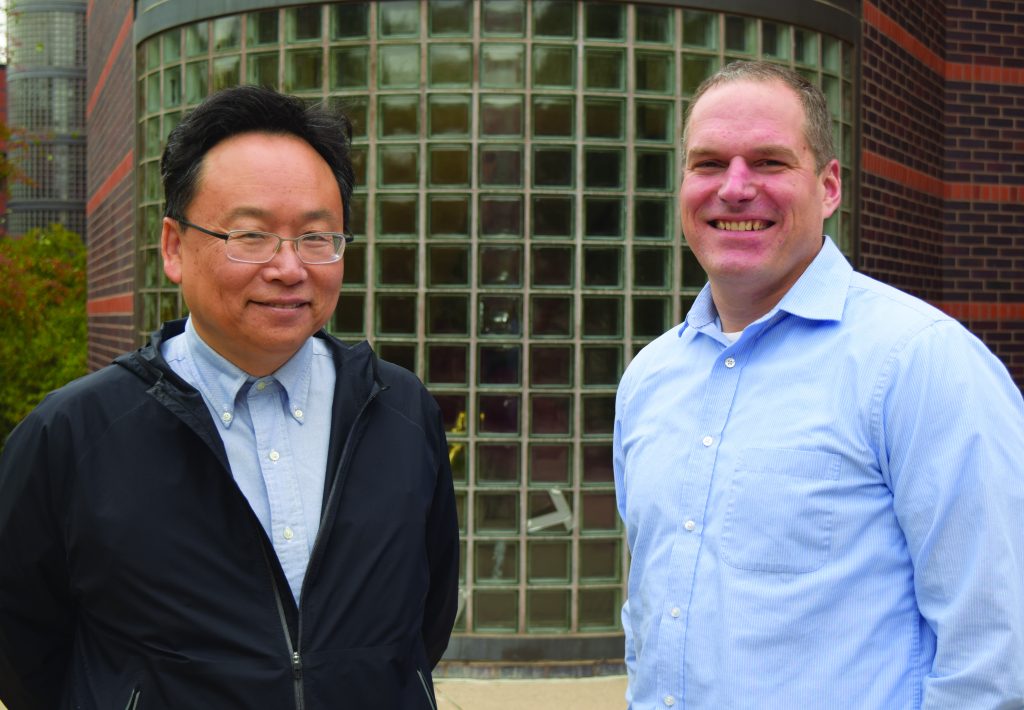 Industrial engineering associate professor Jo Min poses with industrial engineering assistant professor Cameron MacKenzie. Both men are wearing light blue collared shirts and are posing in front of Black Engineering Building.