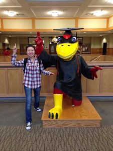 Industrial engineering graduate student Xue Lei poses next to a statue of Cy the Cardinal (mascot for Iowa State University). Cy is wearing a graduation cap and gown, while Xue is wearing jeans and a plaid shirt. 