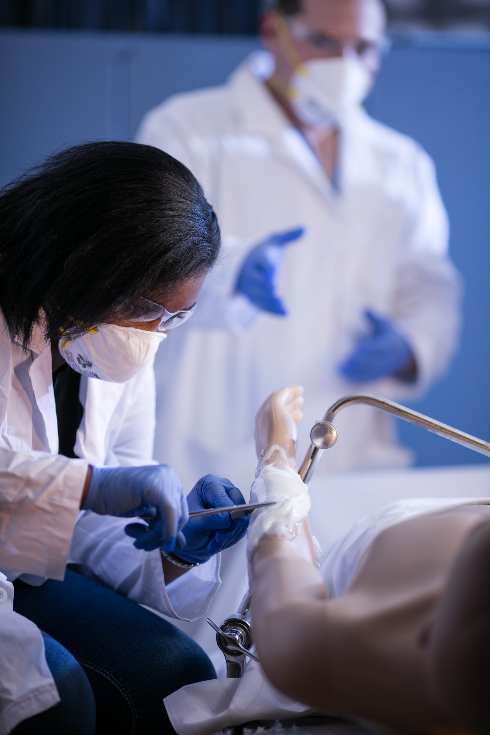 A student performs a surgical operation on a mannequin. The student is wearing safety glasses, a face mask, rubber gloves and a white lab coat. A professor can be seen in the background guiding her through the process.