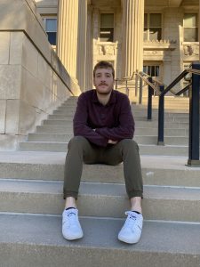 Levi poses on the stairs of a campus building. He's wearing green pants and a maroon shirt.